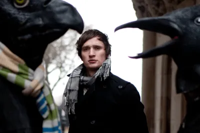 Erland Cooper in a dark coat and scarf between two giant crow-head masks