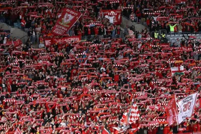 Liverpool FC supporters holding up red-and-white scarves and banners in a packed stadium stand