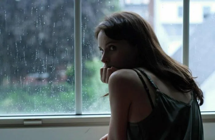 Film still from Breathe In: a young woman looks back from a rain-streaked window