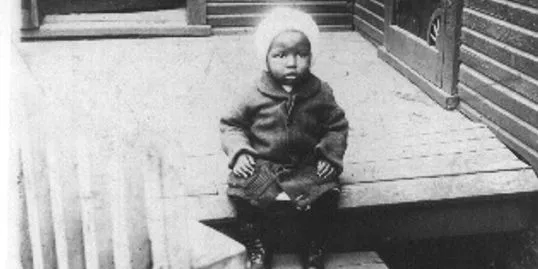 Black-and-white photo of a toddler Billy Strayhorn in a knit cap sitting on a porch