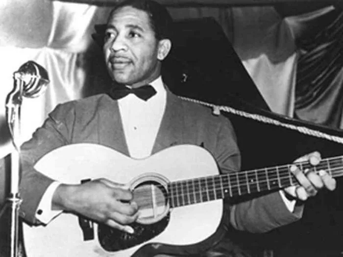 Black-and-white photo of Lonnie Johnson in a bow tie playing acoustic guitar at a microphone