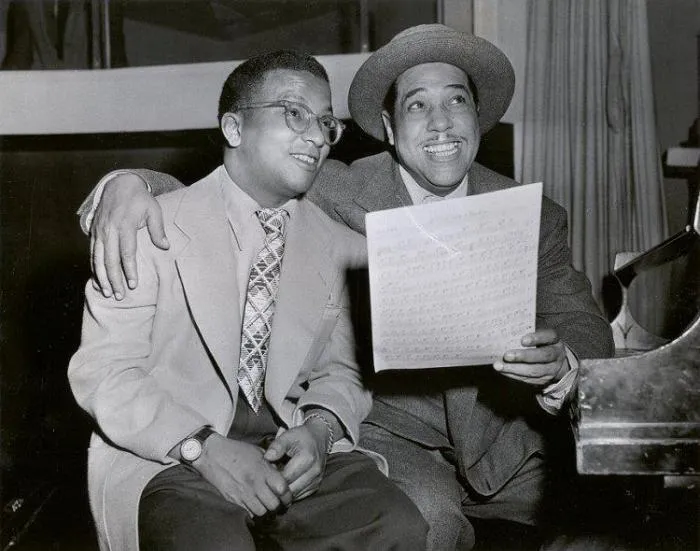Black-and-white photo of Billy Strayhorn and Duke Ellington smiling over sheet music at a piano