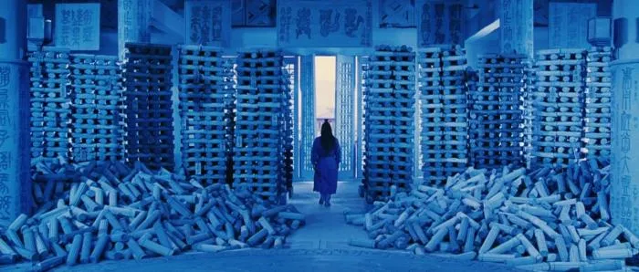 Blue-tinted film still from Hero: a figure walks between towering stacks of bamboo scrolls