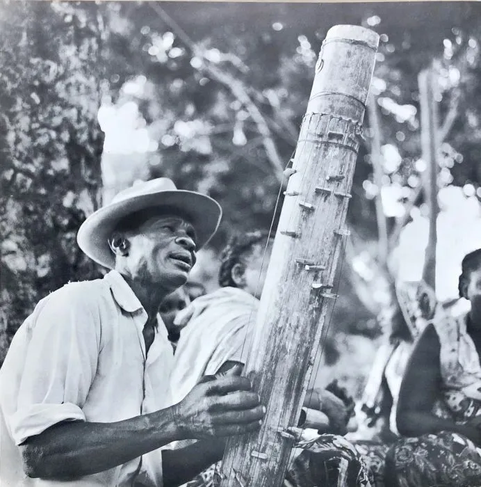 Black-and-white photo of a Malagasy man in a straw hat playing a tubular bamboo valiha